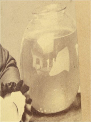 Trimmed mounted studio photograph portrait of a seated woman, with a visible light reflector next to her and a reflected view of the photo studio in a prop fish bowl.