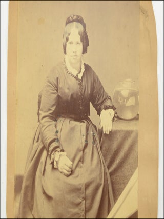 Trimmed mounted studio photograph portrait of a seated woman, with a visible light reflector next to her and a reflected view of the photo studio in a prop fish bowl.